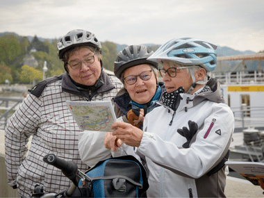 Drei Radfahrer mit Helmen und Brillen studieren eine Karte an einem Hafen an der Donau. Im Hintergrund ist ein weißes Passagierschiff sichtbar.
