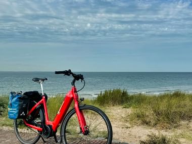 Rotes Elektrofahrrad mit blauen Packtaschen auf Küstenweg in Zeeland, mit Blick auf Nordsee, Dünen und Strandgras unter bewölktem Himmel.