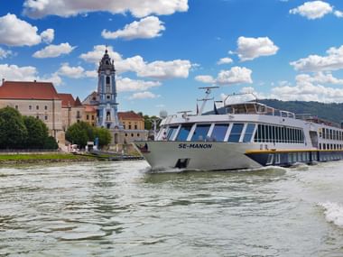 Weißes Kreuzfahrtschiff MS SE-Manon fährt auf Fluss vor historischer österreichischer Stadt mit barockem Kirchturm und mittelalterlichen Gebäuden.