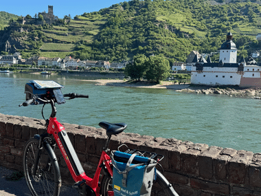 Rotes Fahrrad auf Steinmauer mit Blick auf den Rhein und die Burg Pfalzgrafenstein auf einer Insel. Weinberge und Hügel im Hintergrund.