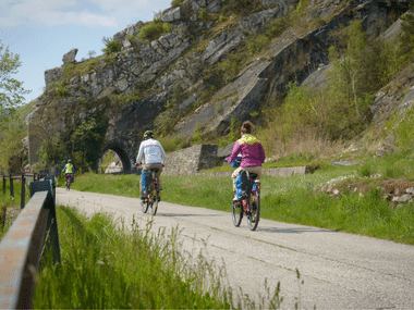 Drei Radfahrer auf einem Schotterweg entlang der Donau, vorbei an einer Felswand mit einem Steinbogentunnel. Grüne Vegetation säumt den Weg.