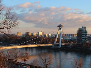 Bridge of the Slovak National Uprising spanning the Danube River in Bratislava at sunset, with modern city skyline and residential buildings.