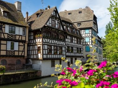 Traditionelle Fachwerkhäuser entlang eines Kanals in Straßburg mit bunten Blumen im Vordergrund und grünen Bäumen unter blauem Himmel.