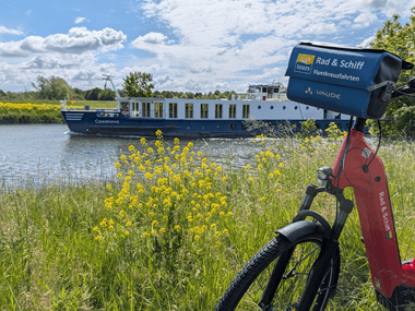 Flusskreuzfahrtschiff Casanova mit blau-weißem Rumpf auf Wasserstraße. Rotes Fahrrad mit Reiseveranstalter-Schild im Vordergrund, gelbe Wildblumen am Ufer.