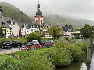 View of Zell with red-white church tower, traditional houses, and vineyard-covered hillsides shrouded in mist. Red rental bikes line the riverbank.