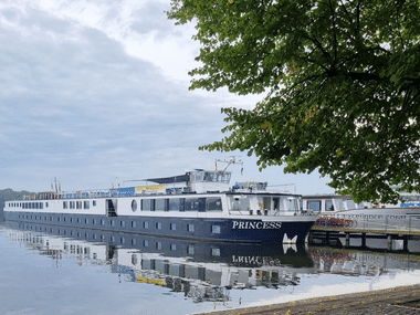 Weiß-blaues Princess Flusskreuzfahrtschiff am Anleger in Berlin. Das Schiff spiegelt sich im ruhigen Wasser, grüne Bäume rahmen die Szene.
