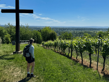 Wanderer mit Rucksack steht bei Holzkreuz in Bodenheim und blickt auf Weinberge und Tallandschaft unter blauem Himmel.