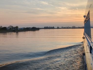 Blick vom Deck der MS SE-MANON bei Sonnenuntergang mit goldenen Reflexionen auf ruhigem Wasser, ferner Küstenlinie und orange-rosa Himmel.