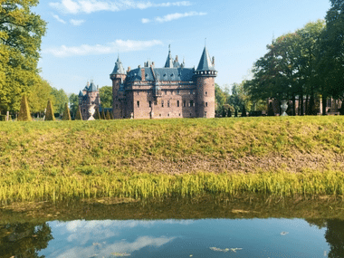 Red brick castle with multiple towers and turrets surrounded by green lawns and trees. A moat reflects the blue sky in the foreground.