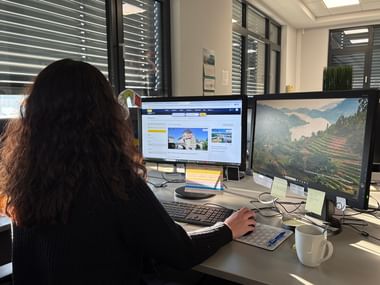 Woman with dark hair sitting at office desk working on dual monitors, one showing travel website, the other a landscape image. Coffee mug nearby.