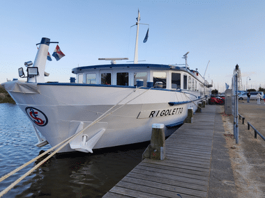 Weißes Flusskreuzfahrtschiff Rigoletto am Holzsteg im Hafen von Enkhuizen. Das Schiff hat eine blaue Flagge und niederländische Farben am Bug.