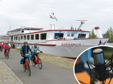 Gruppe von Radfahrern auf einem Uferweg neben dem weißen Flusskreuzfahrtschiff Normandie. Ein Podcast-Mikrofon-Symbol ist eingeblendet.