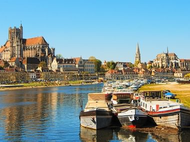 Panoramablick auf Auxerre mit gotischer Kathedrale und historischen Gebäuden entlang der Yonne. Boote am Ufer unter klarem blauen Himmel.