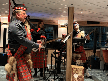 Dudelsackspieler in traditionellem schottischen Kilt und Federmütze spielt drinnen mit Bandmitgliedern an Gitarre und anderen Instrumenten.