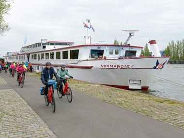 Gruppe von Radfahrern auf einem Kanalweg neben dem Flusskreuzfahrtschiff MS NORMANDIE. Das weiße Schiff mit rotem Rand liegt am Dock.