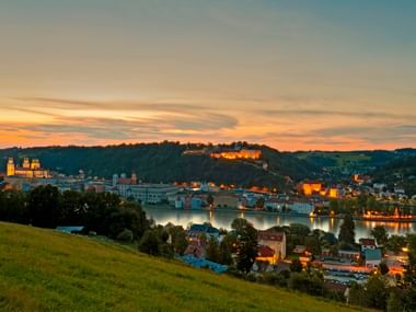 Evening view of Passau with illuminated historic buildings along the Danube River, colorful sunset sky, and Veste Oberhaus fortress on the hill.