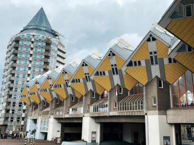 Rotterdam's iconic yellow Cube Houses tilted at 45 degrees next to the cylindrical Pencil Tower under a cloudy sky.