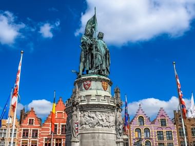Bronzedenkmal mit zwei Figuren und Fahne am Grote Markt in Brügge, umgeben von bunten mittelalterlichen Gildenhäusern unter blauem Himmel.