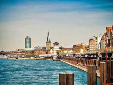 Düsseldorf Rhine waterfront with city skyline View of Düsseldorf's Rhine waterfront with wooden pier posts, historic church spire, modern glass tower, and colorful buildings along the promenade.