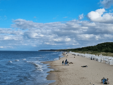 Breiter Sandstrand in Zinnowitz mit weißen Strandkörben, entspannenden Menschen und blauer Ostsee unter bewölktem Himmel. Bewaldete Küste in der Ferne.