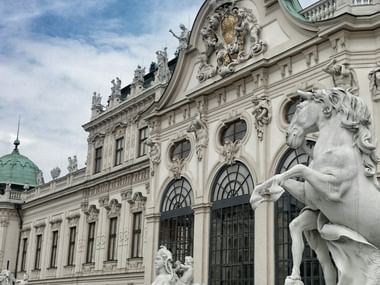 Ornate baroque facade of Belvedere Palace in Vienna with white stone sculptures, arched windows, and decorative elements under a cloudy sky.