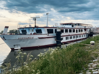 White river cruise ship named Normandie with red trim moored at a cobblestone dock. Green vegetation in foreground, cloudy evening sky above.