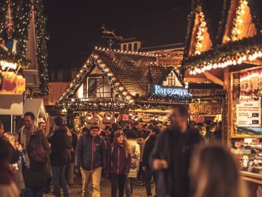 Christmas market with festive lights at night Evening scene at a Christmas market with wooden stalls decorated with warm lights. Visitors walk between illuminated booths selling food and gifts.