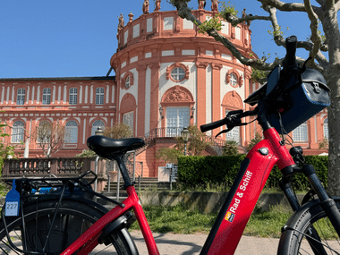 Rotes E-Bike auf Rasen vor dem barocken Schloss Biebrich mit seiner markanten rosa Fassade und prächtiger Architektur unter blauem Himmel.