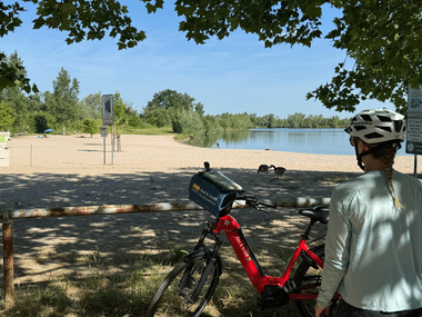 Radfahrer mit rotem Fahrrad und Helm betrachtet Sandstrand am Badesee Eich. Bäume rahmen die Szene, blaues Wasser und grüne Vegetation im Hintergrund.