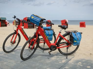 Zwei rote Fahrräder mit blauen Packtaschen auf Holzsteg am Ostseestrand mit bunten Strandkörben im Hintergrund.
