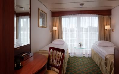 Twin cabin with two single beds, green patterned carpet, glass table, wooden desk, mirror, wall lamps, and large window with beige curtains.
