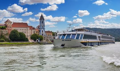 Weißes Kreuzfahrtschiff MS SE-Manon fährt auf Fluss vor historischer österreichischer Stadt mit barockem Kirchturm und mittelalterlichen Gebäuden.