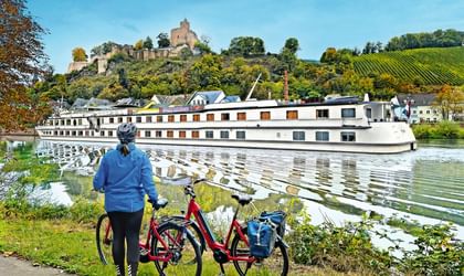 Radfahrer mit rotem Fahrrad beobachtet weißes Flusskreuzfahrtschiff. Burg auf Hügel mit Weinbergen im Hintergrund unter blauem Himmel.