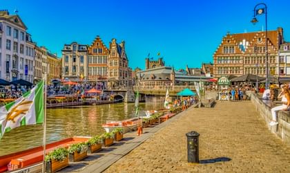 Kopfsteinpflaster-Uferpromenade in Gents Altstadt mit mittelalterlichen Zunfthäusern, Kanalbooten und Touristen unter blauem Himmel.