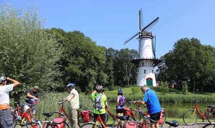 Gruppe von Radfahrern mit roten Fahrrädern vor einer traditionellen weißen holländischen Windmühle umgeben von grünen Bäumen und Wasser.