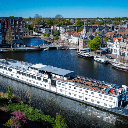 Aerial view of white river cruise ship De Nassau sailing through a Dutch harbor with historic buildings and other boats moored along the waterfront.