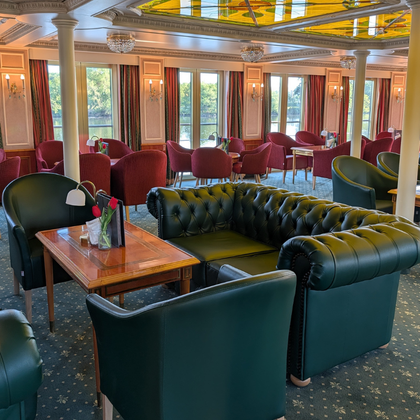 Eleganter Salon an Bord der Casanova mit grünen Chesterfield-Ledersofas, roten Samtstühlen, Holztischen, weißen Säulen und großen Fenstern.