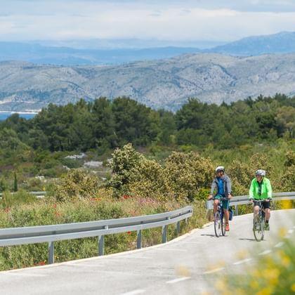 Zwei Radfahrer auf einer asphaltierten Straße bei Hvar mit Bergen, Meer und üppiger Vegetation im Hintergrund unter teilweise bewölktem Himmel.