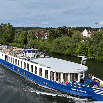 Blue and white river cruise ship Casanova sailing on a river with a green landscape and village with traditional houses in the background.