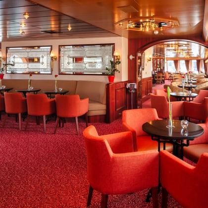 Elegant salon bar on MS Switzerland with red carpeting, orange upholstered chairs, round tables, and decorative mirrors on walls.