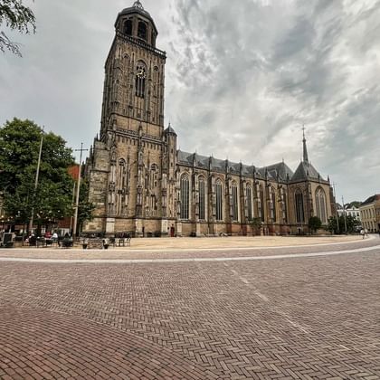 Large Gothic church with prominent stone tower in Deventer's town square, surrounded by cobblestones and historic buildings under cloudy sky.