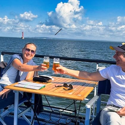 Zwei Passagiere stoßen mit Biergläsern an einem Tisch auf einer Fähre über das Stettiner Haff an, mit blauem Himmel, Wolken und Wasser im Hintergrund.