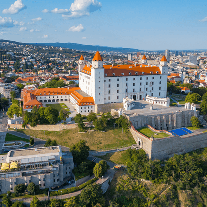 Aerial view of Bratislava Castle with white walls and orange roofs on a hilltop, surrounded by the city and green landscape under blue sky.