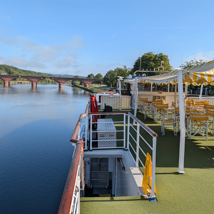 Sonnendeck des Flusskreuzfahrtschiffs Casanova mit gelb-weiß gestreiftem Sonnensegel, Holzstühlen und grünem Kunstrasen. Fluss und Brücke im Hintergrund.