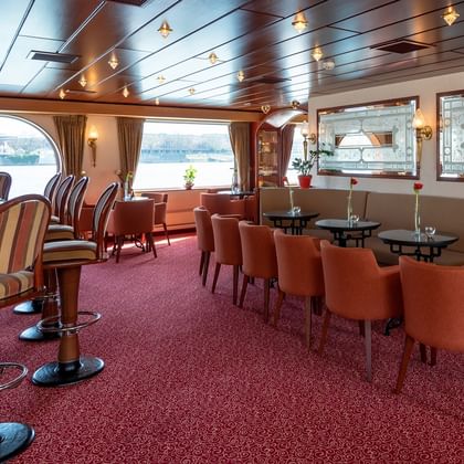 Elegante Salon-Bar mit orangefarbenen Stühlen, runden Tischen, Barhockern und großen Fenstern mit Blick aufs Wasser. Roter Teppich und Holzverkleidung.