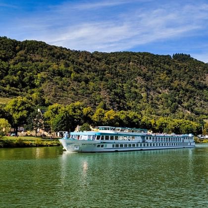 Weißes Flusskreuzfahrtschiff MS Switzerland fährt auf ruhigem grünen Wasser mit bewaldeten Hügeln und Dorfgebäuden im Hintergrund unter blauem Himmel.