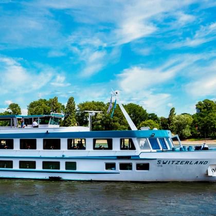 White river cruise ship MS Switzerland sailing on calm water with blue sky, trees and greenery visible along the shore in the background.