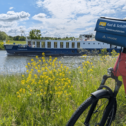 Flusskreuzfahrtschiff Casanova mit blau-weißem Rumpf auf Wasserstraße. Rotes Fahrrad mit Reiseveranstalter-Schild im Vordergrund, gelbe Wildblumen am Ufer.
