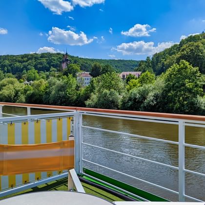 Sonnendeck der MS Casanova mit gelben und orangen Sitzgelegenheiten, weißen Geländern mit Blick auf Fluss mit bewaldeten Hügeln und Kirchturm.