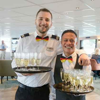 Zwei lächelnde Besatzungsmitglieder der MS Princess in Uniform mit Tabletts voller Champagnergläser im Schiffslounge-Bereich mit Passagieren im Hintergrund.
